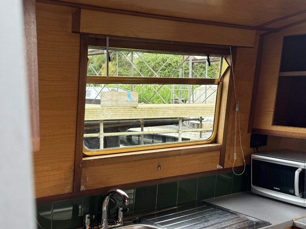 An interior view of a wooden cabin with a window showing a dock outside. The window is partially open, revealing green surroundings and a structure in the background. A stainless steel sink and a microwave are visible in the kitchen area below the window.