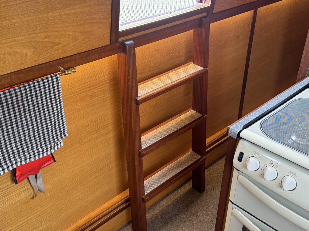 A wooden ladder with four steps is positioned next to a stove in a kitchen. The wall behind features light-colored wooden panels, and a black and white checkered towel is hanging nearby.
