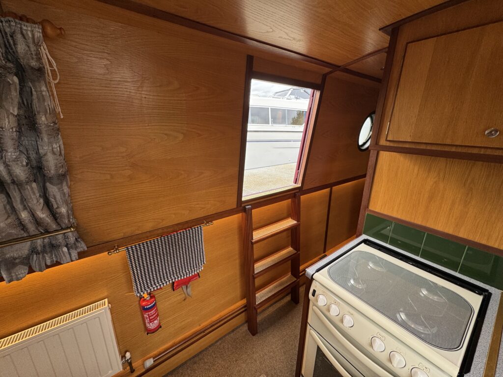The interior of a boat kitchen features wooden walls and cabinetry with a small round window. A white stove is positioned next to a sink, with a towel hanging on a rack nearby. Shelves are visible to the left, and there is a heater on the wall.
