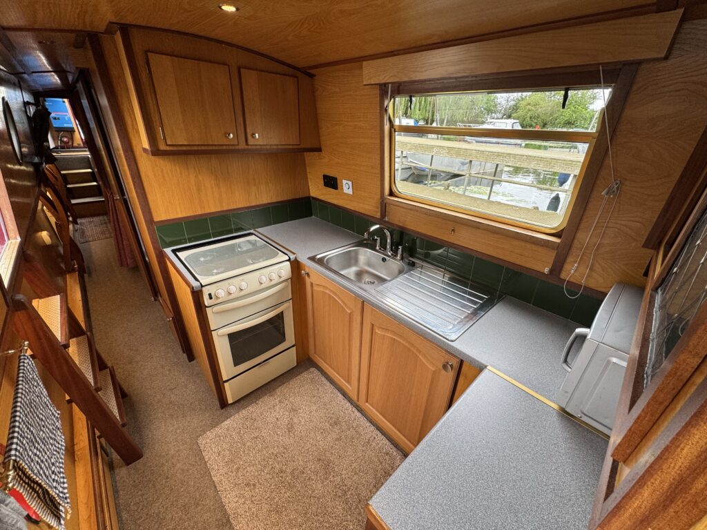 An interior view of a narrowboat kitchen featuring wooden cabinetry and a stainless steel sink. The kitchen includes a gas stove and a countertop with a towel hanging nearby. A window provides natural light, with greenery visible outside.