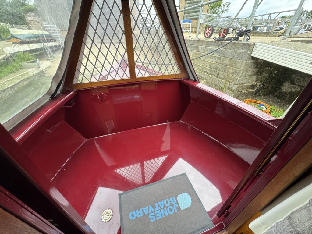 An interior view of a boat featuring a maroon-colored hull with glossy surfaces. A rubber mat with the logo "Jones Boatyard" is visible on the floor. Natural light enters through a diamond-patterned window, revealing equipment and structures outside.
