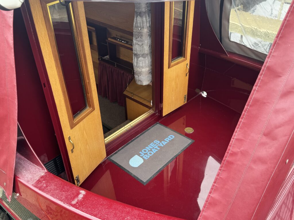 A view of the interior of a boat with open double doors revealing wooden cabinetry. The floor has a glossy red surface, and a mat with the text 'Jones Boatyard' is placed by the entrance. The background shows soft furnishings and a window.