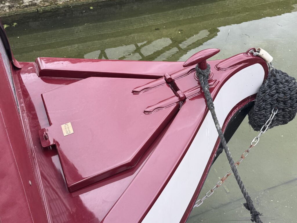 The close-up image shows the bow of a red and white boat, featuring a triangular hatch that is securely fastened with metal latches. A thick rope is coiled nearby, and the water reflects the boat’s colors.