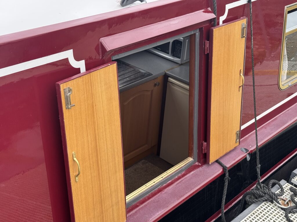 A maroon narrowboat features a window with two wooden foldable doors opened, revealing a glimpse of the interior kitchen area with tan cabinets and a countertop. The boat's exterior is decorated with a white design along the side.