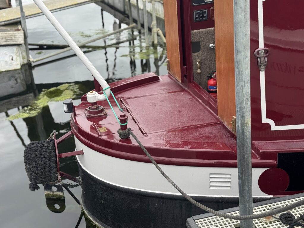 A close-up view of the stern of a burgundy boat docked at a marina. The boat features a fender and various equipment, with reflections visible on the water surface. The background includes wooden dock structures and green aquatic plants.
