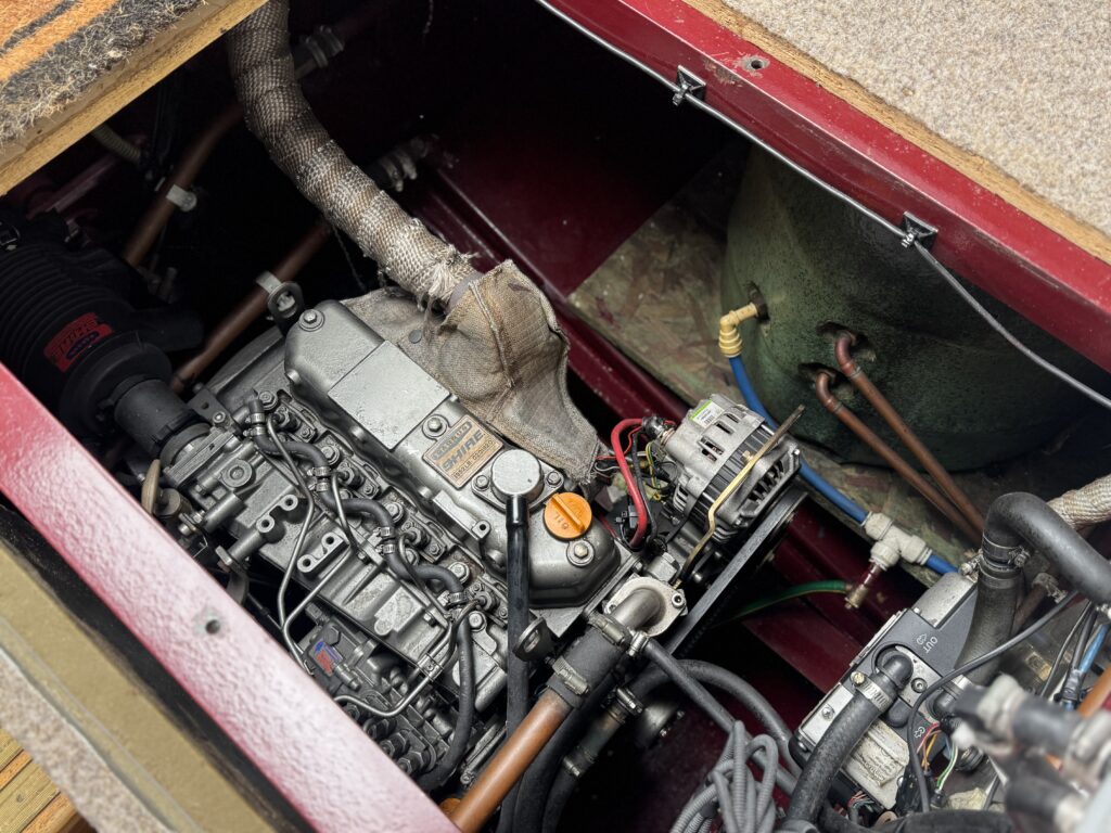 An overhead view of a mechanical engine compartment showing various components, including a metal engine block, hoses, wires, and a green cylindrical tank. The area is characterized by a mix of metallic and rubber materials.