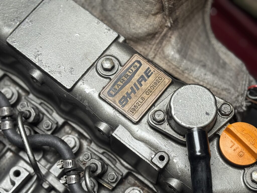 A close-up image of an engine component featuring a silver metal surface with the engraved logo "HKS" and the word "SHINE". There are various metal pipes and connections, and a circular cap labeled "OIL" in orange on the right.