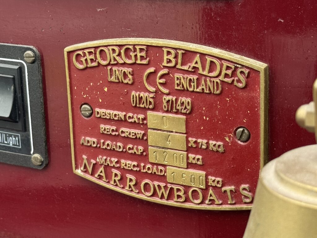 A red metal plate with gold lettering and engraved information mounted on a surface. The plate includes the name "George Blades" along with registration numbers and specifications for a narrowboat, detailing design category, crew capacity, added load capacity, and maximum load.