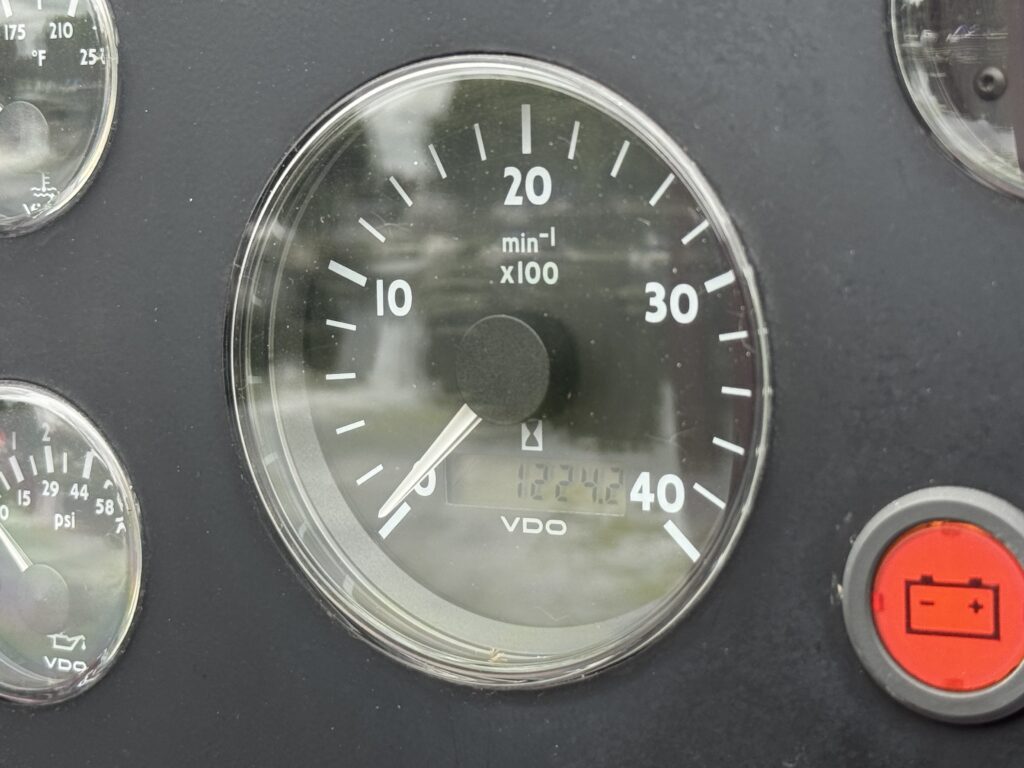 A close-up view of a tachometer displaying engine RPM. The gauge shows a range from 0 to 40, with the needle pointing to 30. Other gauges are visible in the background, along with a red button and a battery symbol.