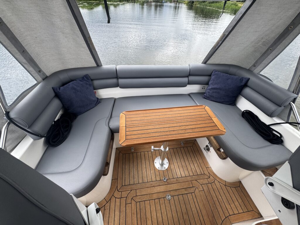 A boat interior featuring a curved seating area with gray cushions and blue throw pillows. A wooden table sits in the center, and the floor is made of polished wooden planks. Outside, the water is visible with greenery nearby.