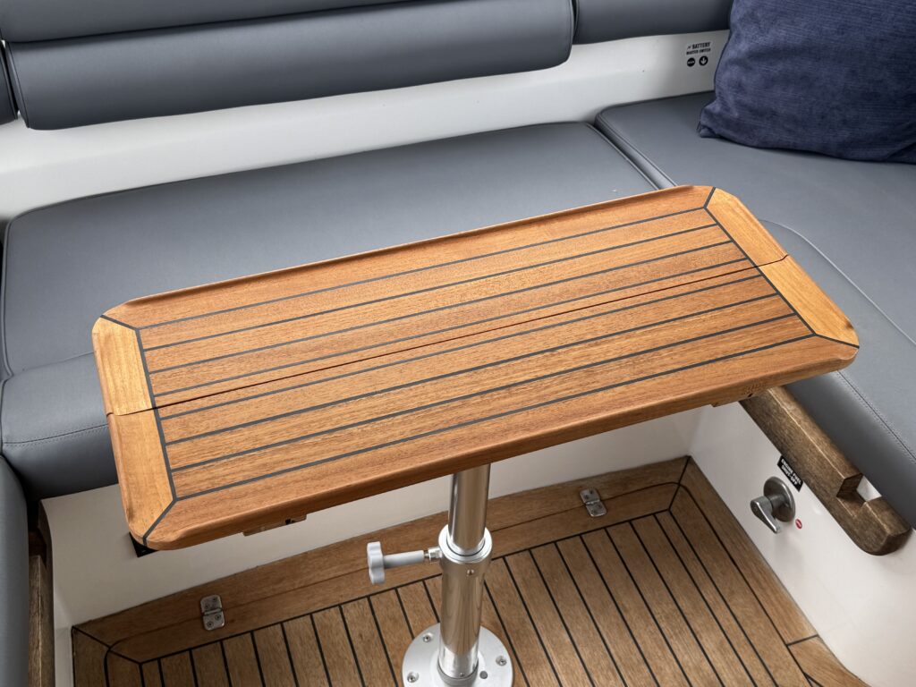 A wooden table with horizontal slats is mounted on a polished metal pole, situated on a boat. The surrounding seating area features gray cushions and a dark blue pillow, with a teak wood floor visible below.