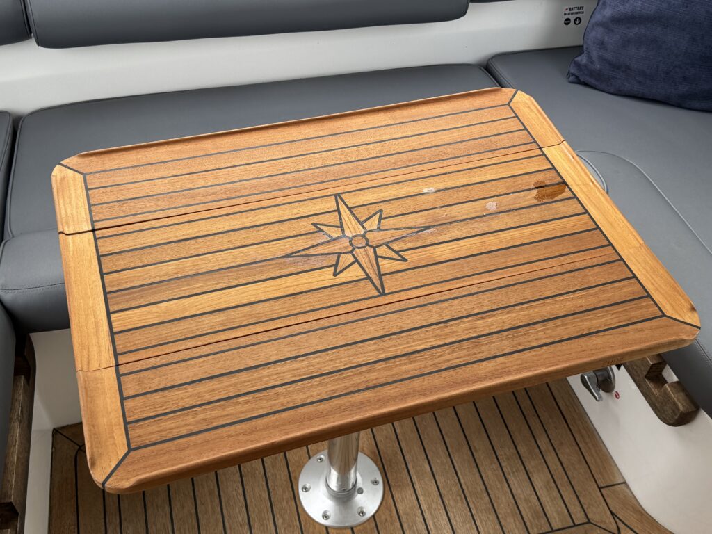 A wooden table with a compass design in the center sits on a boat. The table has a polished surface with dark lines forming a layered pattern and is supported by a round metallic pedestal, surrounded by cushioned seating.