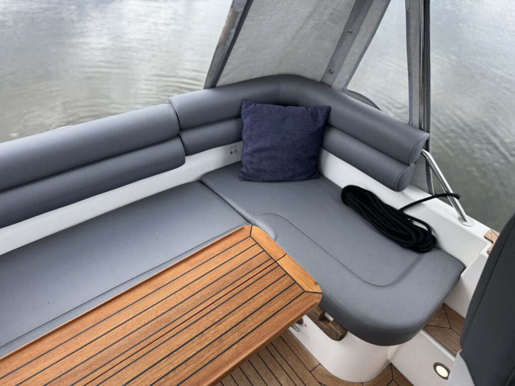 A sectional seating area on a boat featuring gray cushions and a dark blue pillow. A wooden table is positioned beside the seating, with a rope laid out next to it. The background shows calm water and a partly cloudy sky.