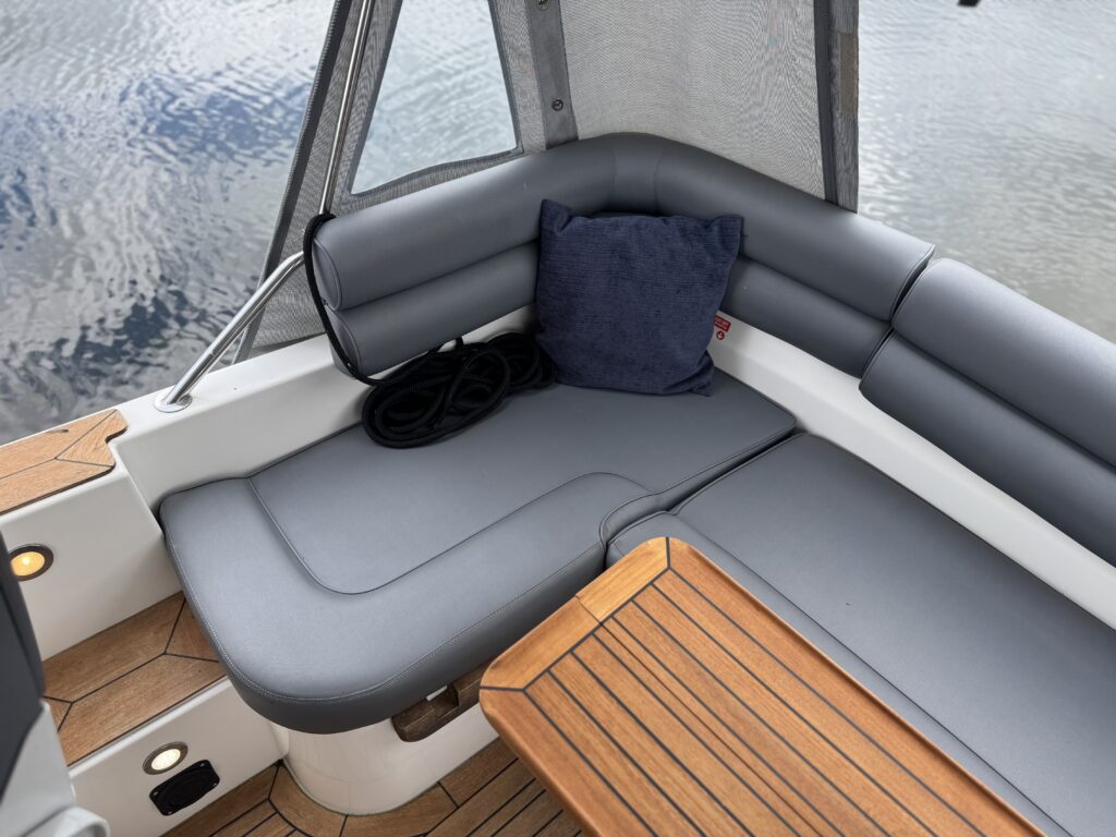 A corner seating area on a boat featuring gray cushioned seating. A dark blue pillow is placed on the seat, and a wooden table with slatted design is positioned nearby. The background shows water reflecting soft ripples.