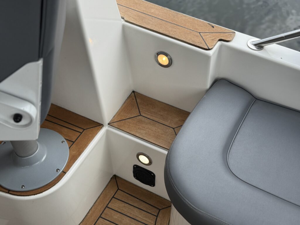 A close-up view of a boat's interior showing a gray seat and light wooden flooring with geometric patterns. A polished metal chair base is visible, along with round lights embedded in the walls. The background reflects water.