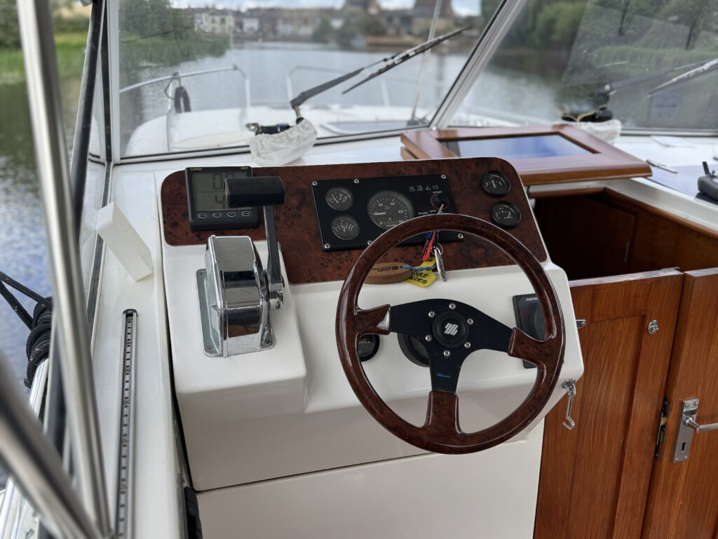 A boat's dashboard featuring a wooden steering wheel and various gauges, including speed and fuel level indicators. The console also includes a throttle control and a small wooden shelf, with a view of water and trees through the front windows.