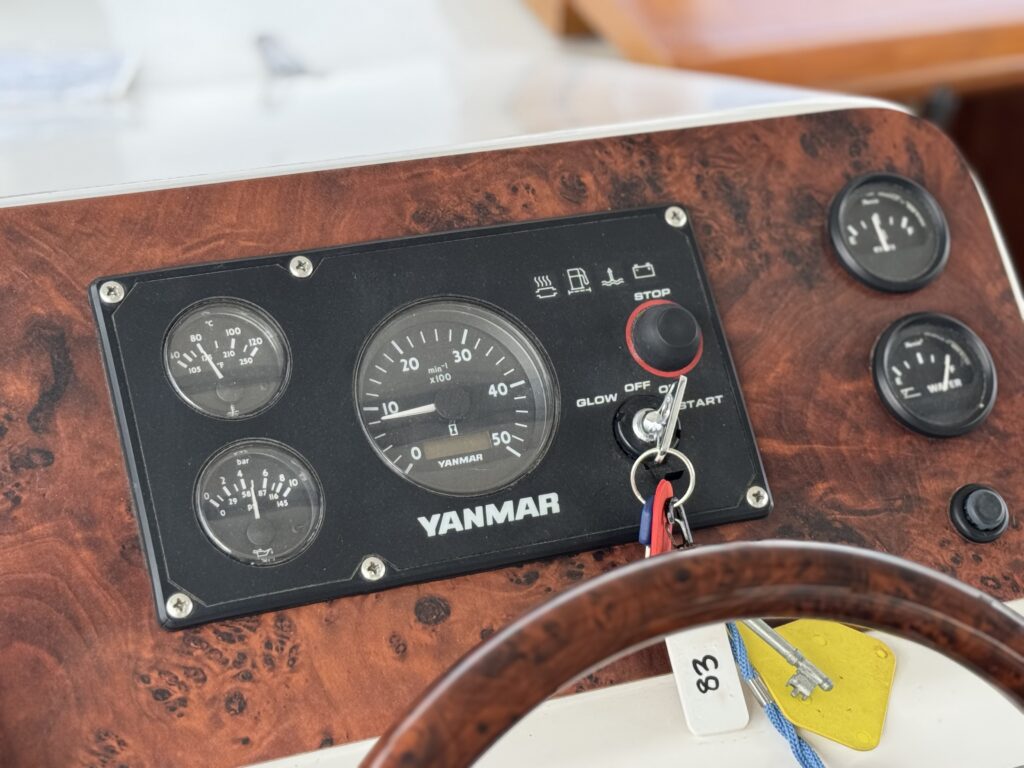 A boat control panel featuring a series of gauges, including a tachometer, oil pressure gauge, and fuel gauge, all beneath a Yanmar logo. The panel is made of wood laminate with a steering wheel partially visible in the foreground.