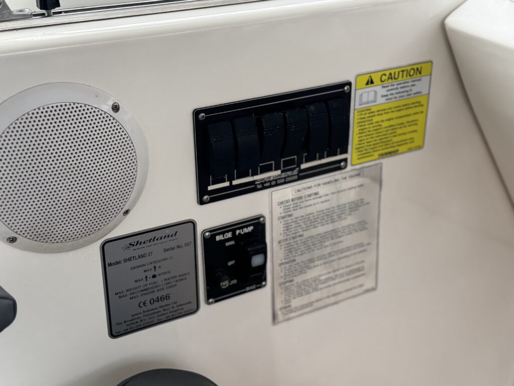 A control panel inside a boat featuring a speaker, a series of toggle switches, a caution sign, and instructional labels. The panel includes a slide pump button and identification tags affixed to a white surface.