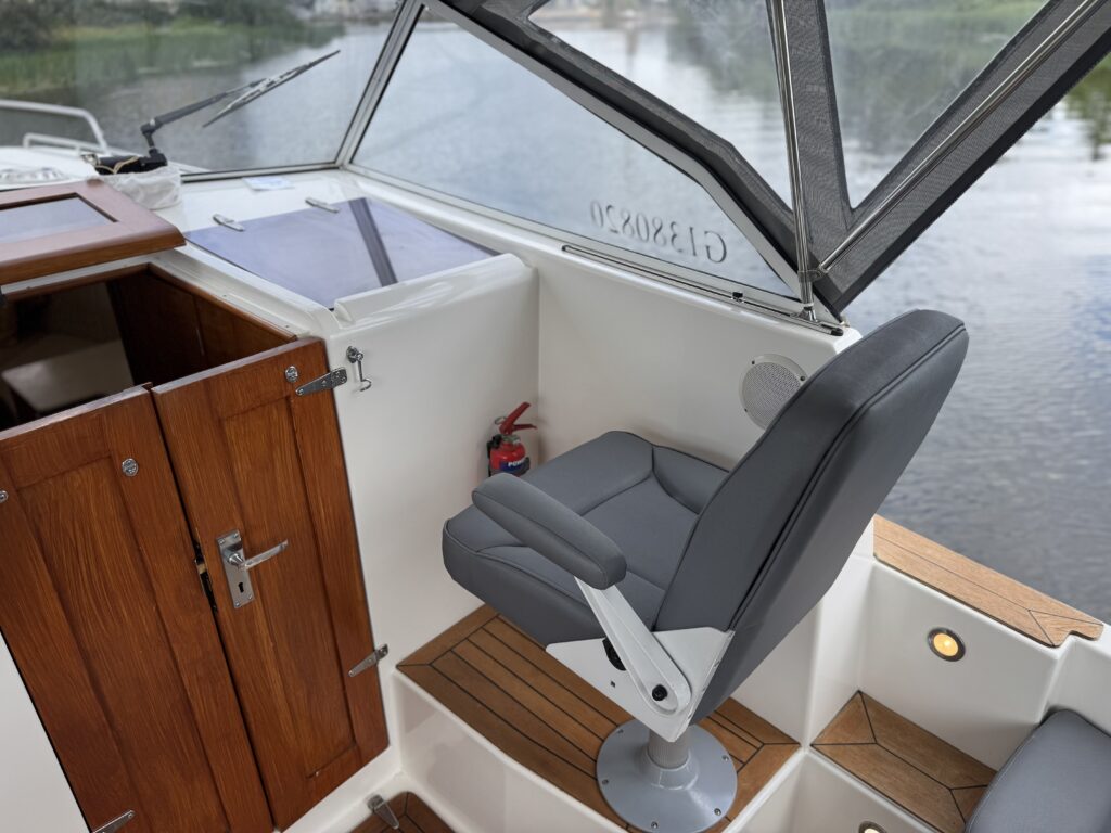The interior of a boat shows a grey swivel captain's chair adjacent to wooden cabinetry. Large windows allow natural light to illuminate the space, and a wooden floor is visible below the seat.
