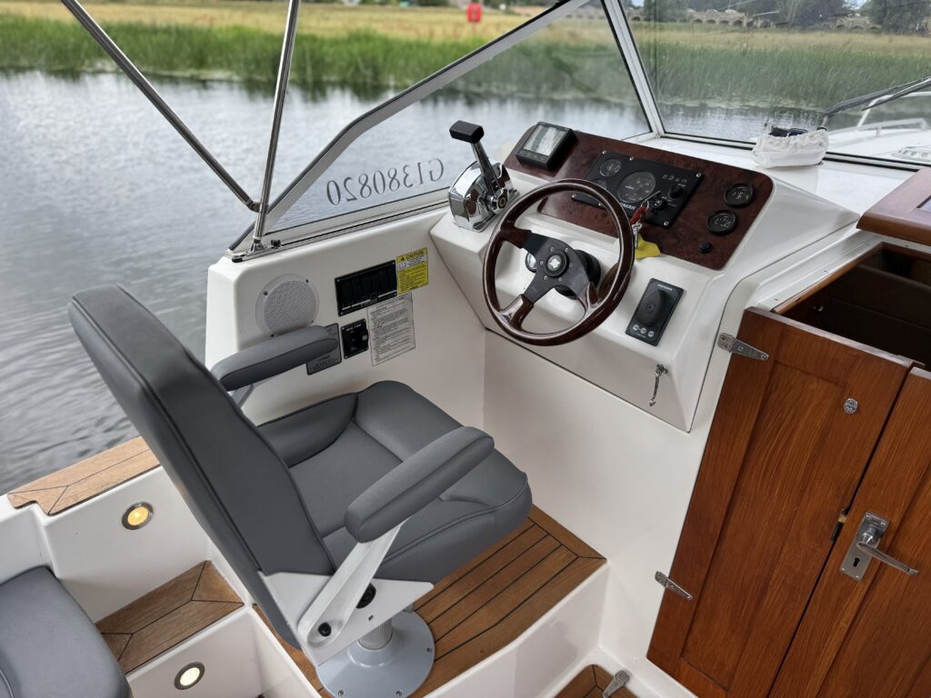 The interior of a boat featuring a gray captain's chair positioned near the steering console. The console includes a wooden dashboard with gauges and controls, while the background shows water and tall grass outside.