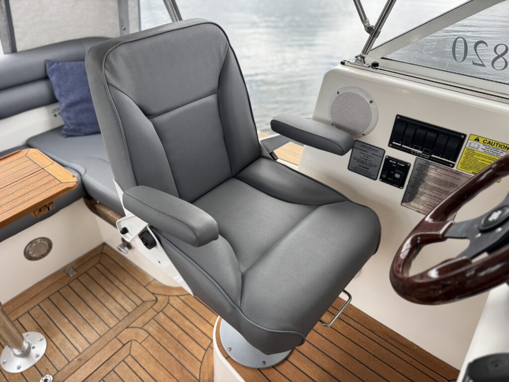 A gray upholstered captain's chair mounted on a boat with armrests, positioned next to a wooden table. The boat interior features a wooden floor and is illuminated by natural light from the nearby window.