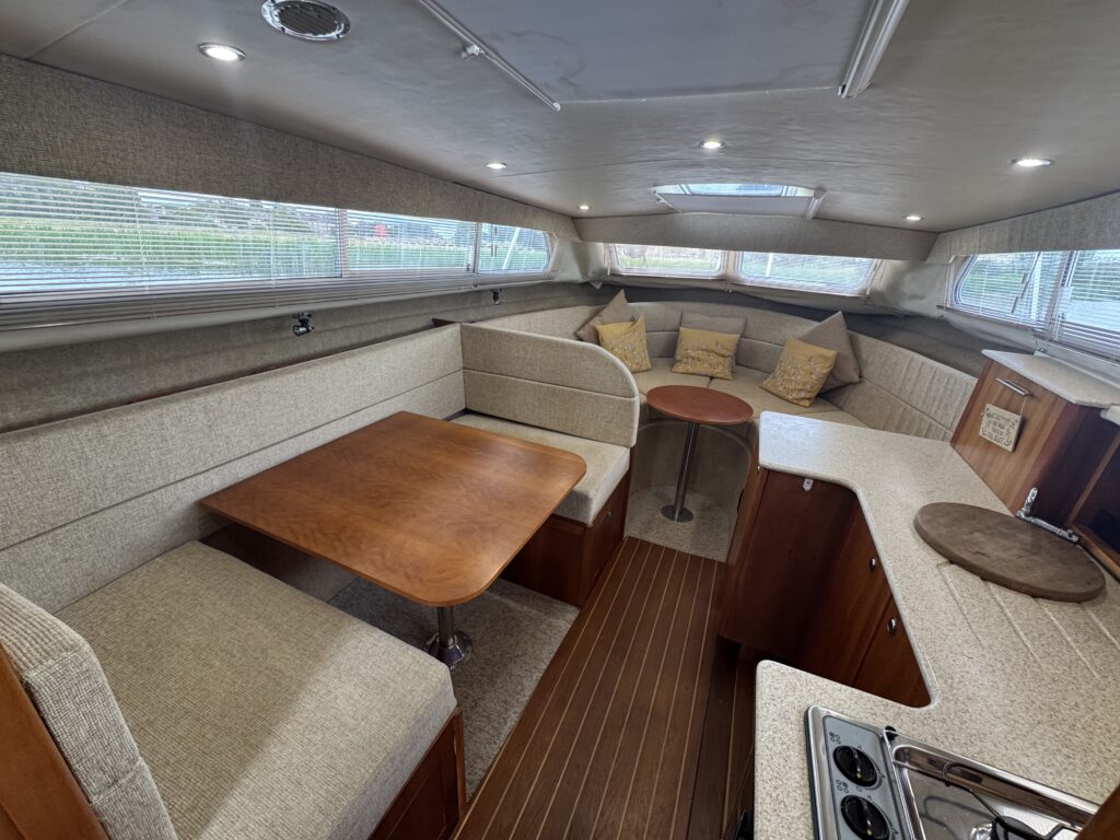 Interior of a sleek boat featuring a cozy seating area with a wooden table, a small circular table, and several cushions. The cabin is well-lit with natural light from large windows, showcasing beige upholstery and wooden accents.