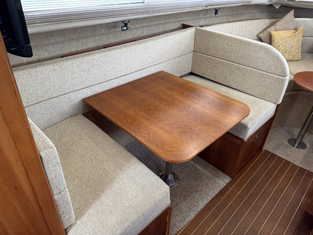 A small dining area on a boat featuring a polished wooden table with a round base and cushioned bench seats upholstered in light fabric. The backdrop includes beige walls and a decorative pillow on one side.