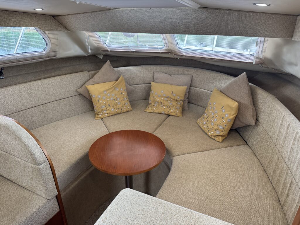 An interior seating area of a boat features a curved beige sofa with comfortable cushions, including two yellow decorative pillows. A small round wooden table is positioned in the center, illuminated by natural light through the windows.