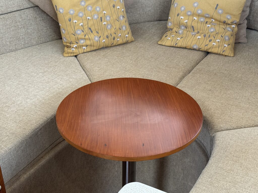 A round wooden table with a smooth surface positioned between a corner couch and two decorative cushions with floral patterns. The cushions are placed against a light fabric background.