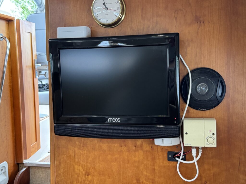 A black flat-screen television mounted on a wooden wall with a clock above it. To the right, there is a round speaker, and below the TV, a small white device is attached to the wall, along with visible wires.