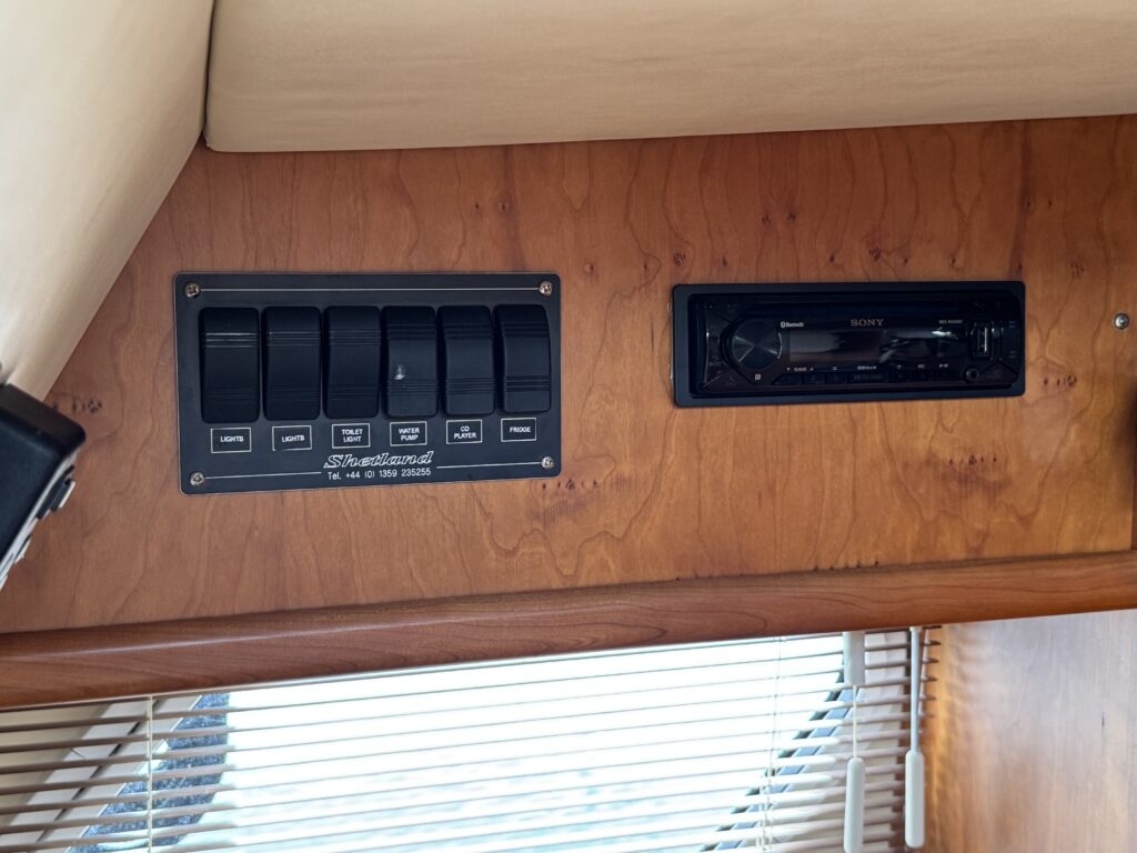 A wooden panel in a marine environment featuring a row of six black switches with labeled functions. Adjacent is a round marine stereo system, both mounted above a wooden counter with light reflections from nearby windows.
