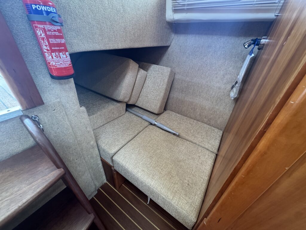 An interior view of a boat featuring a corner seating area with beige cushions arranged against the wall. The area includes a fire extinguisher mounted on the wall and wooden steps leading to the exterior. Light enters from a nearby window.