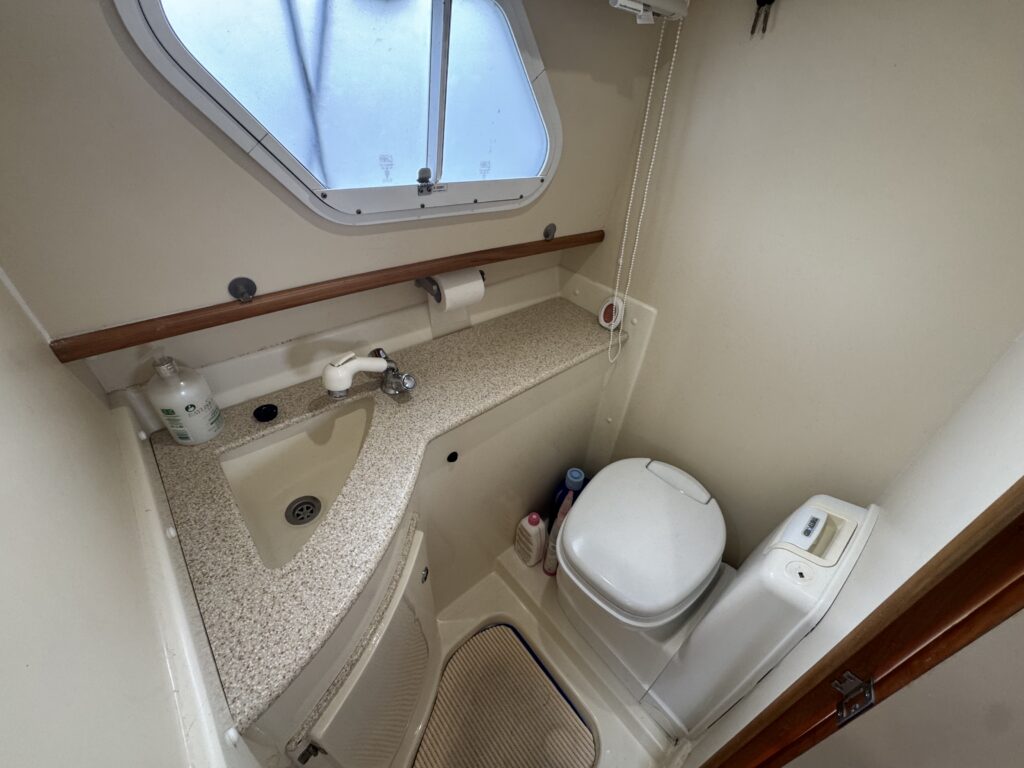 A compact bathroom on a boat, featuring a sink with a faucet, a toilet, and a small window for natural light. The countertop is white with speckles, and a tan mat is placed on the floor. Accessories like soap and toiletries are visible.