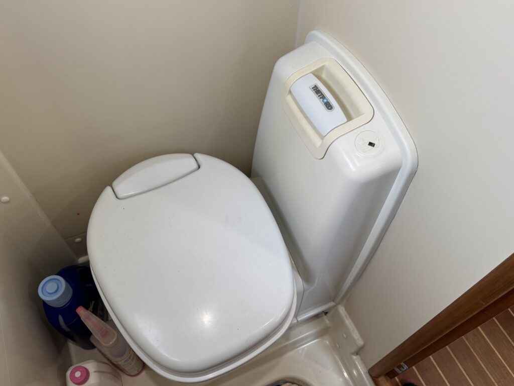 A compact white toilet with a closed lid, positioned in a small bathroom. The wall beside it shows a smooth surface, and various cleaning supplies are visible on the floor and shelf nearby.