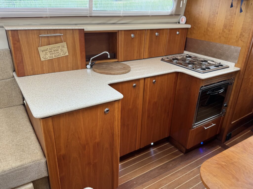A compact kitchen area in a boat featuring wooden cabinetry with multiple drawers and compartments, a countertop with a circular cutting board, a gas stove, and an oven. The space has natural light from a window above the sink.