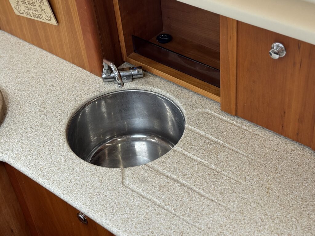 A stainless steel sink set into a textured light-colored countertop. The countertop features linear grooves next to the sink and is bordered by wooden cabinetry. A faucet is mounted above the sink, with a small space for water drainage.