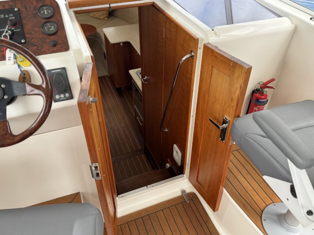 An interior view of a boat showing a wooden door leading to a lower cabin area. The doorway is framed by polished wooden panels, with stairs visible and a white cushioned seat at the bottom. Nearby, there is a gray captain's chair and a control panel with gauges.
