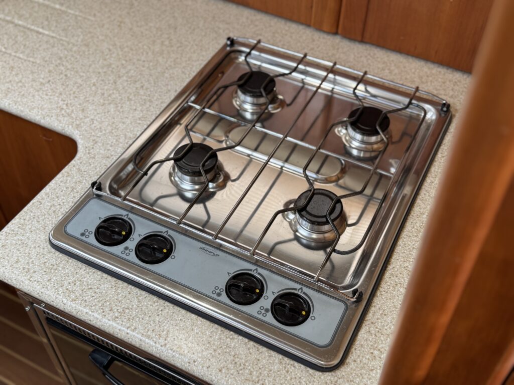 A stainless steel gas stovetop with four burners, each with a black knob, installed on a beige countertop. The background includes wooden cabinetry, emphasizing a compact kitchen design.