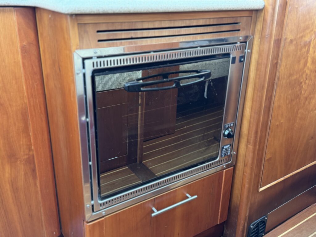 A built-in oven with a glass front and metal frame is integrated into wooden cabinetry. The oven features a handle and digital controls on the right side, with a drawer located beneath for storage.