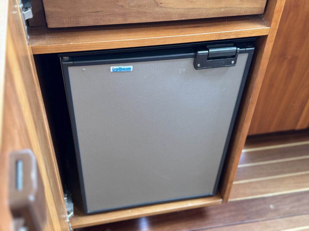 A compact, gray refrigerator with a black door is installed in a wooden cabinet. The refrigerator features a small handle and a label indicating the brand, positioned within a well-crafted kitchen space with wooden cabinetry.