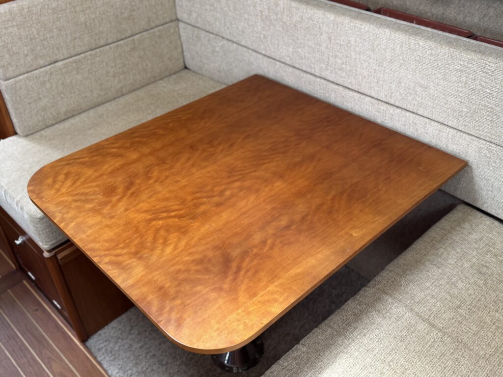 A polished wooden dining table with rounded corners is placed between two gray upholstered benches in a boat's seating area. The table has a rich, warm finish, and the surrounding cushions are light gray.