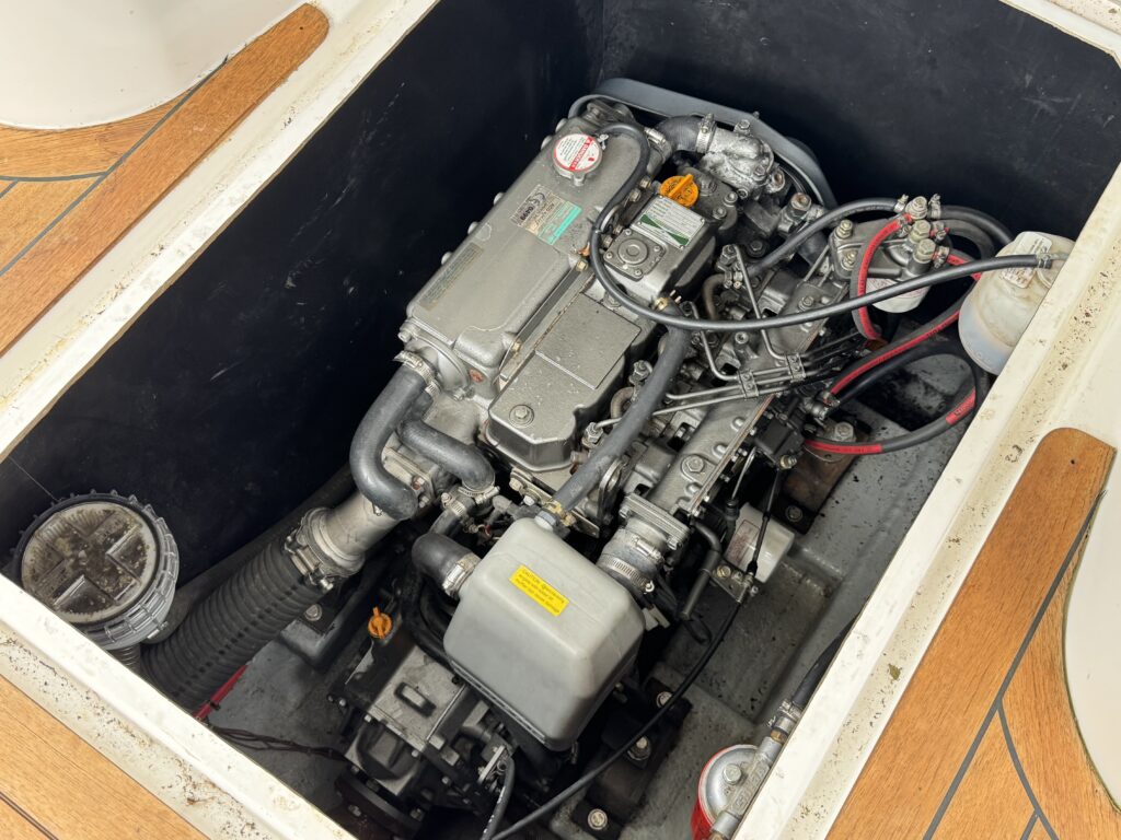 A boat engine compartment showing a marine engine with various pipes and wires. The engine is mounted on a silver base, surrounded by a dark panel and wooden flooring, with additional components visible at the side.