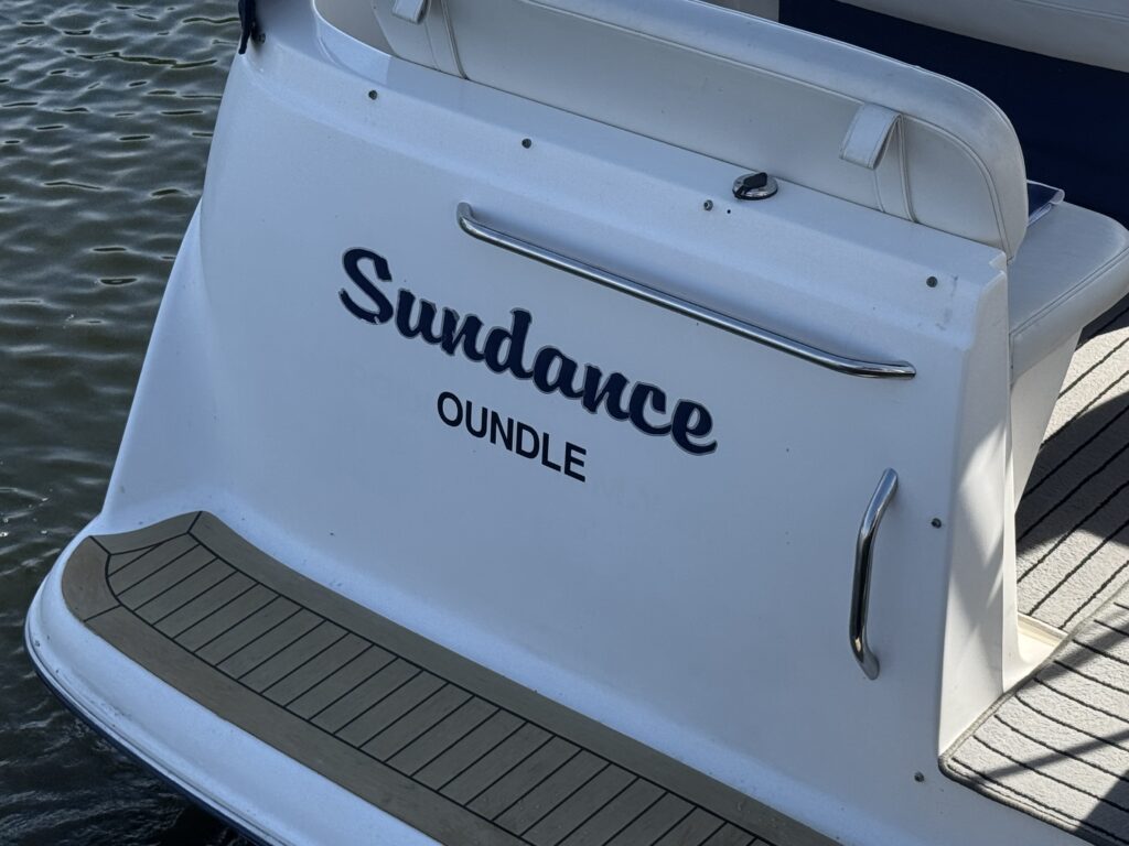 The rear of a white boat features the name "Sundance" in large dark lettering above the word "OUNDLE" in smaller text. The boat's deck has a wooden finish, and water is visible in the background.