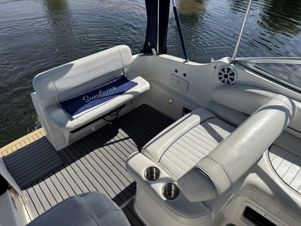 An interior view of a boat featuring light gray seating and a blue cushioned seat with the name "SunLounge". The flooring is a dark gray textured surface, and the background shows calm water.