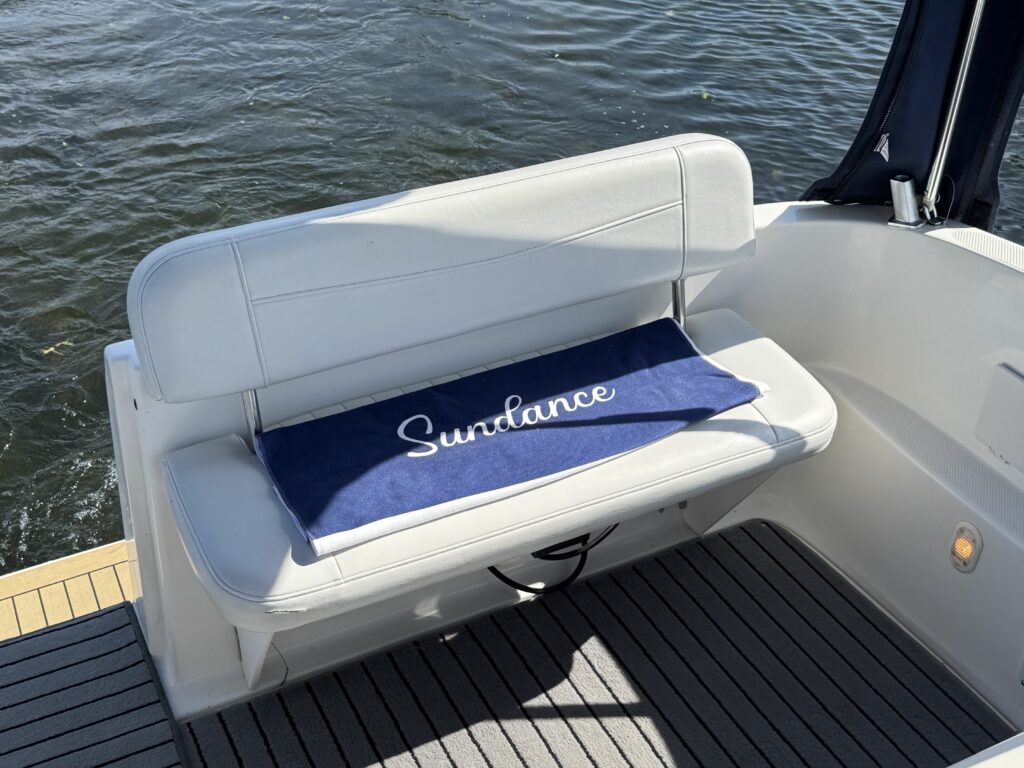 A white cushioned boat seat is positioned on a wooden deck, facing water. A navy blue towel with the word "Sundance" embroidered in white is placed on the seat, with rippling water visible in the background.