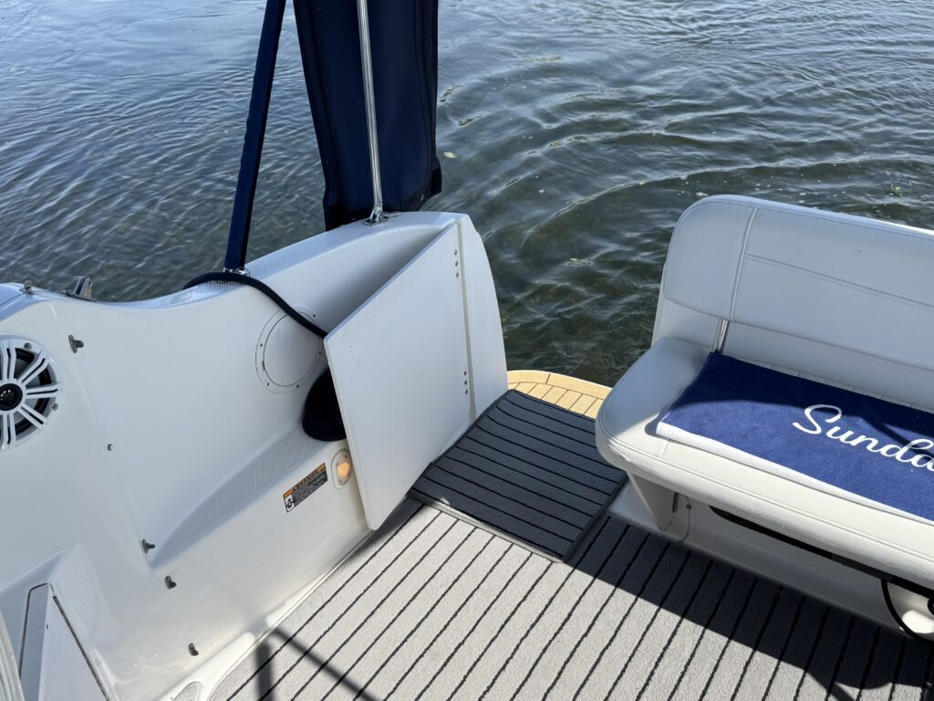 A view of the interior of a boat showing part of a white seat with blue cushion labeled 'Sundancer' and a section of the deck with gray flooring. A door is visible that leads to the storage compartment, with water and reflections in the background.