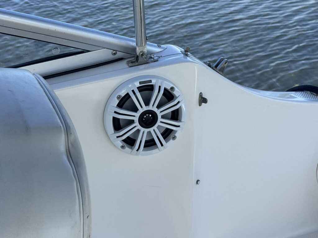 A white marine speaker mounted on the side of a boat, with a circular design featuring slats and a black central opening. The background shows water reflecting light, indicating a sunny day.