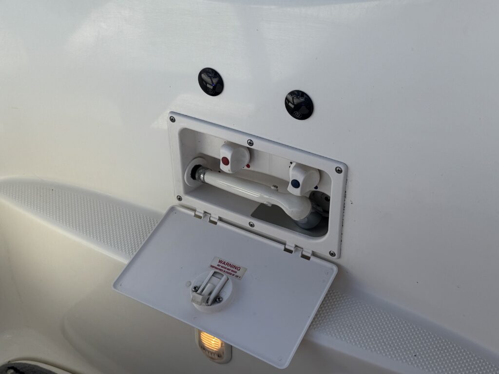 A white exterior panel featuring a small, opened compartment with a pipe and two colored connectors, one red and one blue, visible inside. Two black circular covers are mounted above the compartment, with a small light visible below.