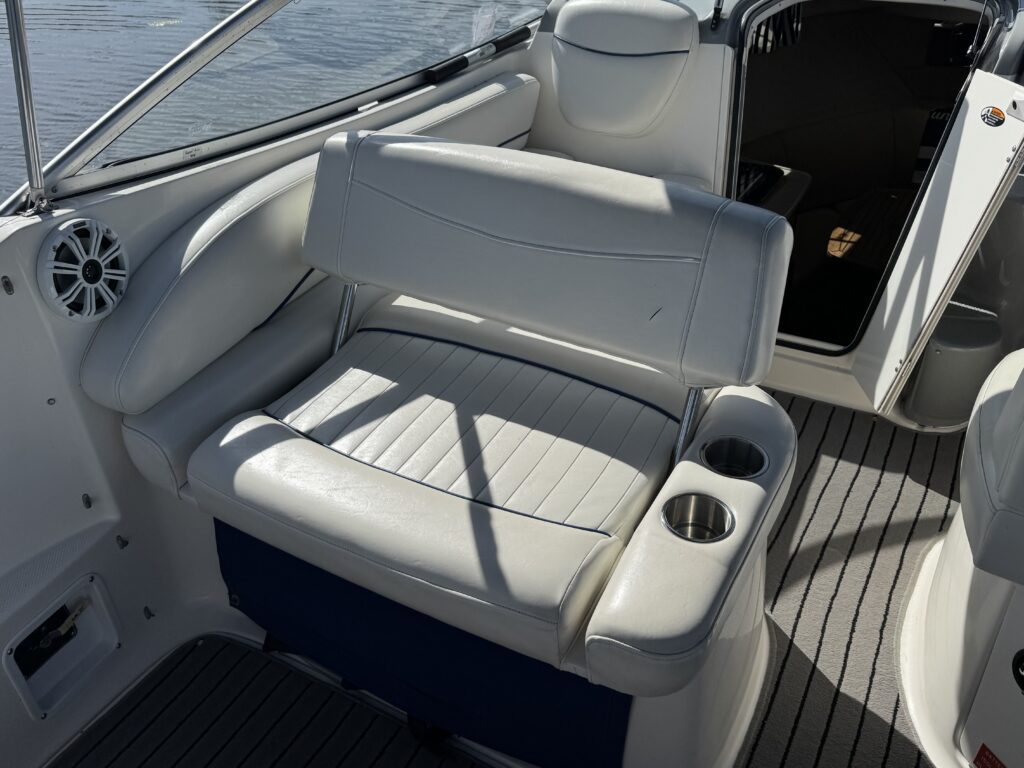 A white vinyl bench seat with a padded backrest and armrest, located on a boat. The seat is positioned near a large window, and there are cup holders on the side. The boat's interior features a gray striped floor and a doorway leading to another compartment.