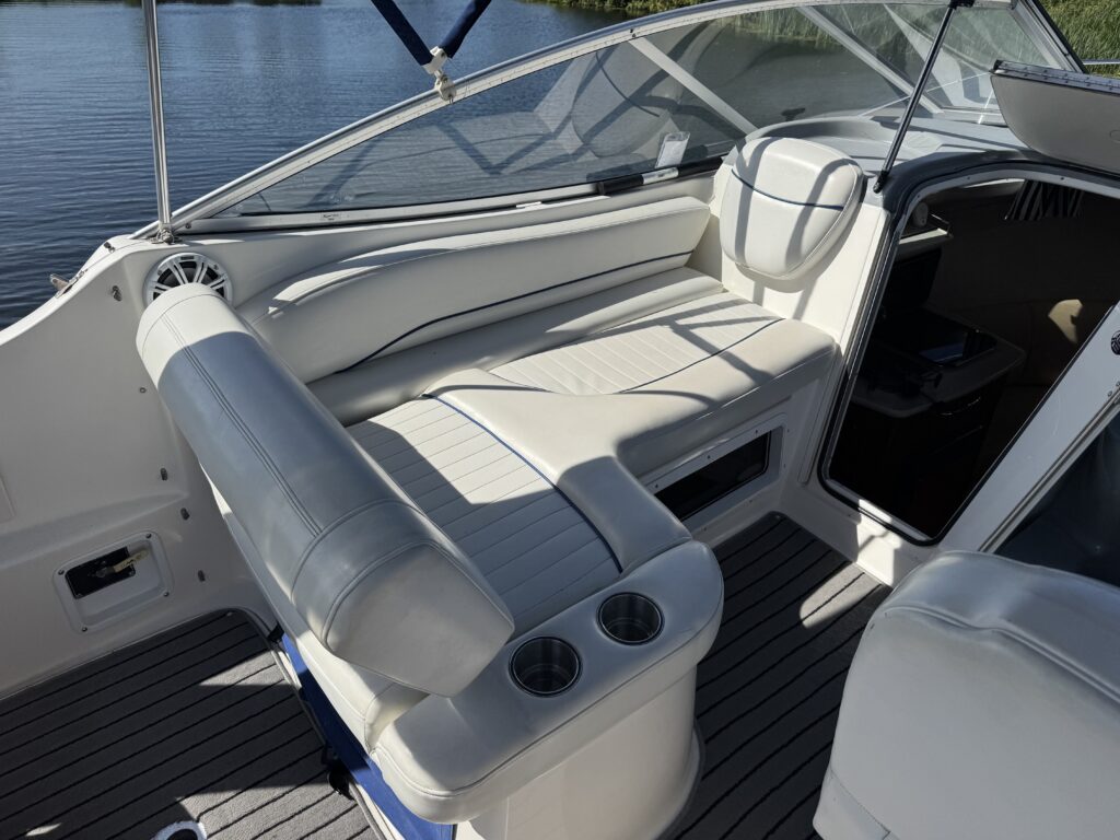 A spacious boat interior featuring white vinyl seating with a bench and two cup holders. The setting is illuminated by natural light, with a view of calm water in the background.